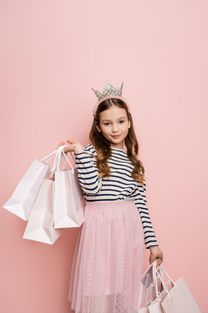 Carefree preteen kid in crown headband holding shopping bags on pink backgroundの写真素材