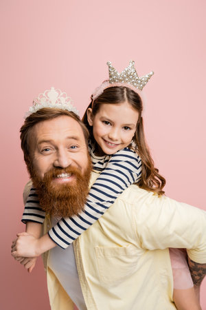 Positive girl piggybacking on father with crown headband isolated on pinkの写真素材