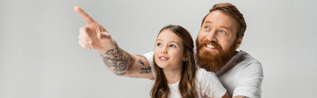 Positive tattooed dad pointing with finger near preteen daughter isolated on grey, bannerの写真素材