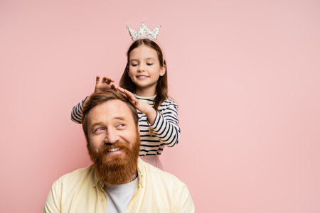 Cheerful girl in crown headband touching hair of smiling dad isolated on pinkの写真素材