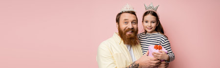 Positive man and daughter with crown headbands holding gift isolated on pink, bannerの写真素材