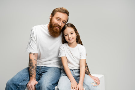 Positive tattooed man sitting near daughter on cube isolated on greyの写真素材