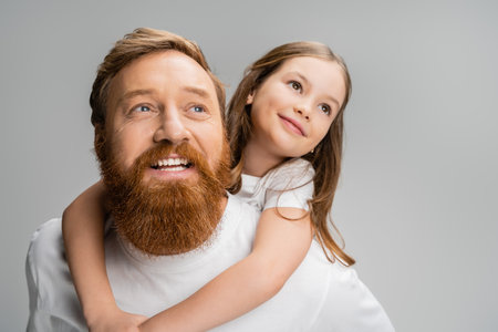 Preteen kid looking away while piggybacking on smiling bearded dad isolated on greyの写真素材