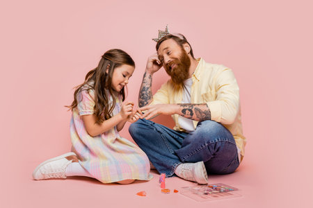 Cheerful girl applying nail polish on hand of tattooed dad with crown headband on pink backgroundの写真素材