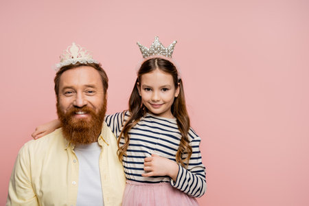 Smiling daughter and father in crown headbands looking at camera isolated on pinkの写真素材