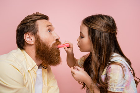 Side view of preteen girl applying lipstick on bearded dad isolated on pinkの写真素材