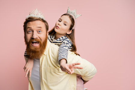 Preteen kid in crown headband showing peace sign while piggybacking on dad isolated on pinkの写真素材