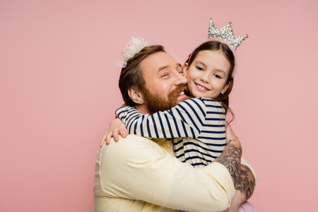 Smiling tattooed father hugging daughter in crown isolated on pinkの写真素材