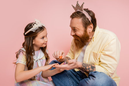 Cheerful man with crown on head holding nail polish near preteen daughter on pink backgroundの写真素材
