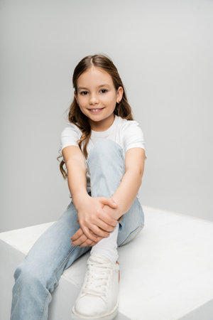 Carefree girl in jeans and t-shirt sitting on white cube isolated on greyの写真素材
