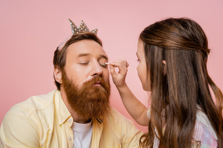 Preteen girl applying eyeshadow on father with crown headband isolated on pinkの写真素材