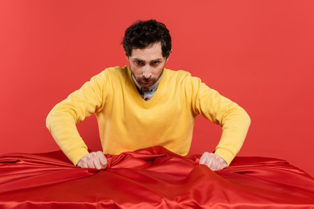 stressed man in yellow jumper pulling red tablecloth on desk isolated on coral backgroundの写真素材