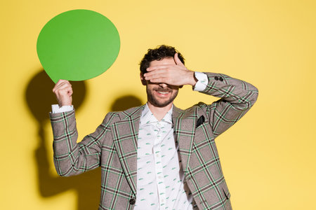 Smiling model covering eyes while holding speech bubble on yellow backgroundの写真素材