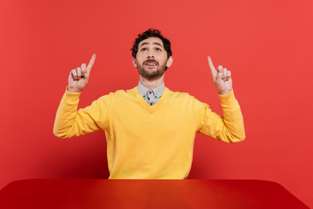 happy man in yellow long sleeve jumper pointing with fingers and looking up on red coral backgroundの写真素材