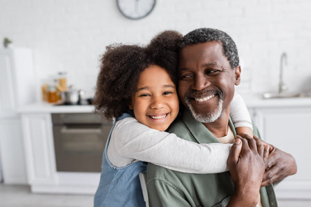 portrait of happy african american girl with curly hair smiling and embracing cheerful grandfather at homeの写真素材