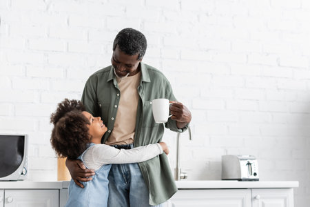 happy african american kid hugging grandfather holding cup with coffee in kitchenの写真素材