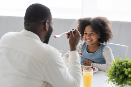 happy african american girl having breakfast and looking at father eating strawberry on blurred foregroundの写真素材