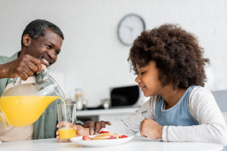 happy african american girl looking at glass while grandfather pouring orange juice during breakfastの写真素材