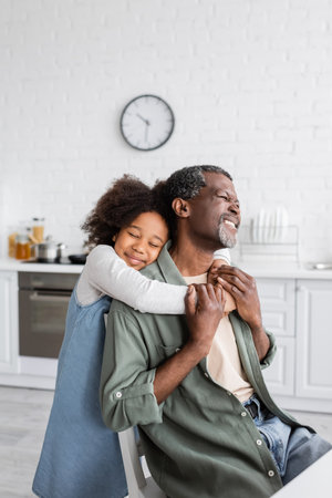 happy african american girl with curly hair smiling and embracing pleased grandfather at homeの写真素材