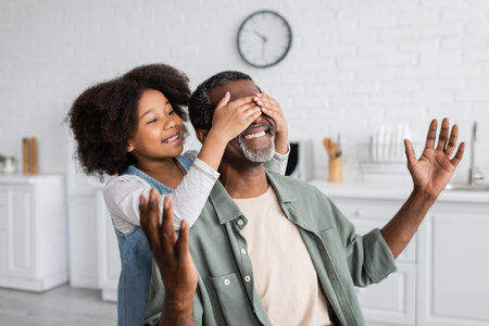 happy african american girl with curly hair covering eyes of smiling grandfather at homeの写真素材