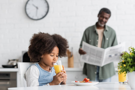 curly african american girl drinking orange juice while grandfather reading newspaper in kitchenの写真素材