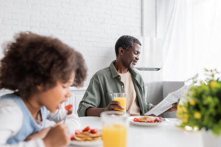 happy african american grandparent reading newspaper while granddaughter having breakfast in kitchenの写真素材