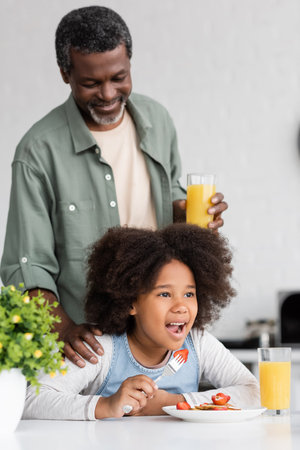 happy african american grandparent holding glass of orange juice and standing behind granddaughter during breakfastの写真素材