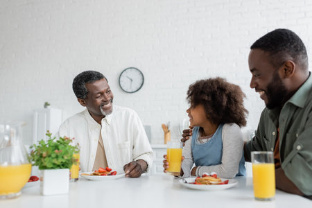 happy middle aged man looking at african american granddaughter near son during family breakfastの写真素材