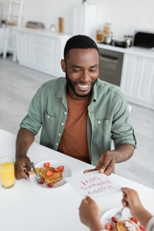 african american girl giving greeting card with happy fathers day lettering to cheerful dad during breakfastの写真素材