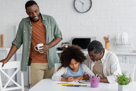 happy african american man holding cup and looking at daughter drawing on paper near his fatherの写真素材