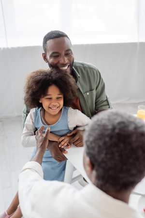 happy african american man hugging curly daughter and looking at father on blurred foregroundの写真素材