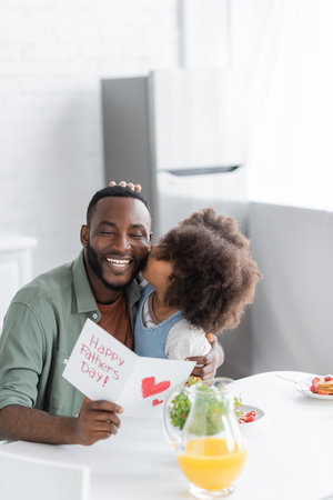 curly african american kid kissing cheek of happy father with greeting card on fathers dayの写真素材