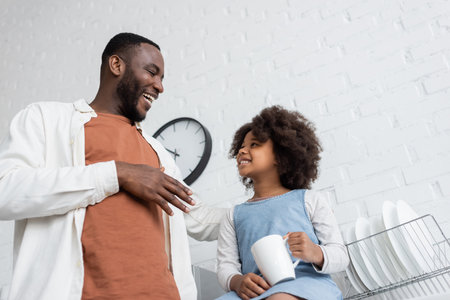 low angle view of joyful african american kid holding cup of tea and looking at happy fatherの写真素材