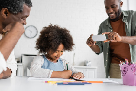 happy african american man taking photo on smartphone while his daughter and father drawing together