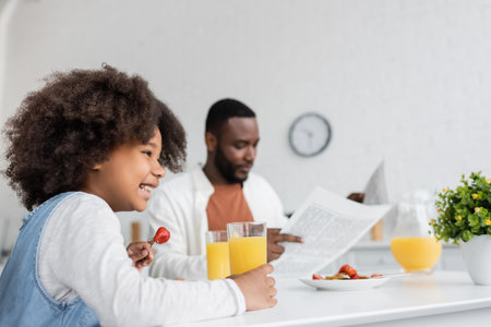 happy african american girl having breakfast while father reading newspaper on blurred backgroundの写真素材