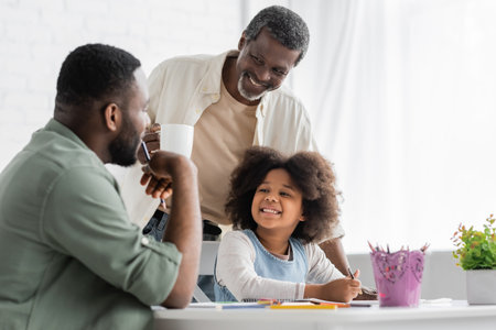 Positive african american child painting and looking at blurred dad near grandfather with coffee at homeの写真素材
