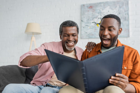 Cheerful and excited african american father and son looking at photo album at homeの写真素材