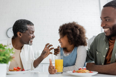 smiling middle aged man looking at african american granddaughter near happy son during family breakfastの写真素材