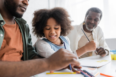 Smiling african american girl painting near father with color pencils and blurred grandpa at homeの写真素材
