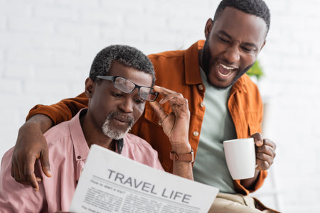 Excited african american man holding coffee cup while father reading newspaper at homeの写真素材