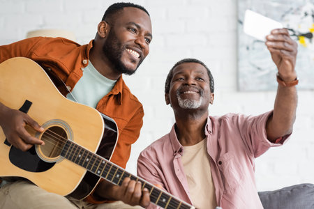 Smiling african american father taking selfie with son playing acoustic guitar at home