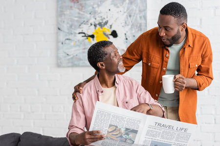 African american man holding cup of coffee and talking to father with newspaper at homeの写真素材