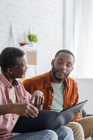 Mature african american man pointing at photo album near son at homeの写真素材