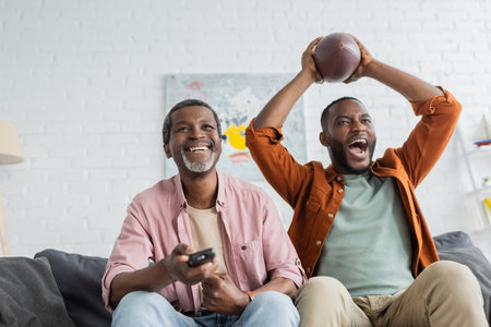 Cheerful african american man holding remote controller near son with rugby ball at home