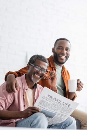 Cheerful african american man holding cup of coffee and hugging mature dad with travel news at homeの写真素材