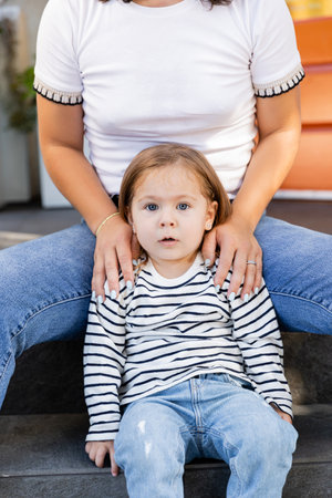 mother in jeans sitting with amazed toddler daughter on porch at backyard against houseの写真素材