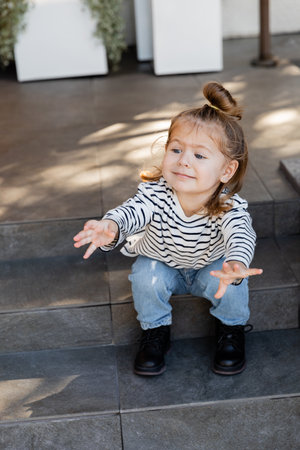 baby girl in casual clothes demanding attention and gesturing while sitting on stairs near houseの写真素材