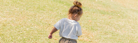 back view of toddler girl in white t-shirt walking on green grass, bannerの写真素材