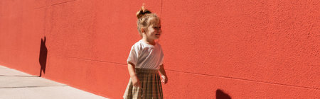 happy toddler girl in checkered skirt and white t-shirt walking near building with red wall, bannerの写真素材