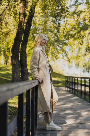 Mature blonde woman in trench coat smiling while standing on bridge in parkの写真素材